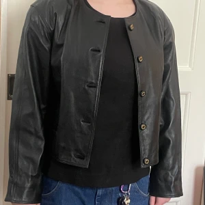 Kort skinnjacka 90s - Välvårdad kort 90-talsjacka i mjukt svart läder, fodrad, med knappar och axelvaddar. 2 små ytterfickor, 1 innerficka. Kemtvätt. Superfint skick! Längd från halslinning fram 63 cm.