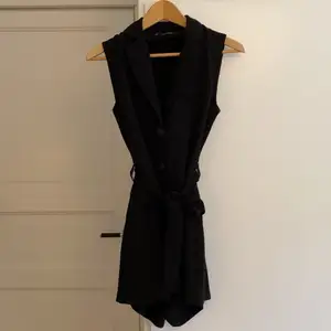En svart klänning med inbyggd byxa, som inte syns vid användning! Blir kaxig med kragen upptill och kroppen får en fin form med tillhörande bälte. Kjolen är delad med ena sidan en ficka och andra sidan plisserad. Väl använd men i gott skick! 
