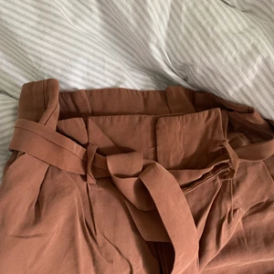 Bruna paper-bag byxor från H&M stlk 38 - Bruna paper-bag byxor från H&M.  Bilden gör dem ej rättvisa - jättefina på!!  Storlek: 38.  Pris: 100 + 66kr. 93% viskos 7% polyester. Fint skick! Endast använda ett fåtal gånger och inget att anmärka på.  Skrynkliga på bild men jag steamar dem såklart innan köpet. 