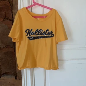 Hollister t-shirt - Croppad t-shirt från hollister, klargul färg med glitterlogga. Strl L men beroende på vad man vill ha för passform kan även en XS bära den. Använd en gång.