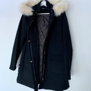 Svart kappa i ullblandning/ helt ny/ stl: 40/ avtagbar päls - Svart kappa i vadderad duffelmodell med två stora fickor med lock framtill. Tillverkad i ullblandning, hög krage samt huva med avtagbar kant i fuskpäls. Kappan är aldrig använd så helt ny. Säljes då den är för stor för mig. Nypris: 1 000 kronor.