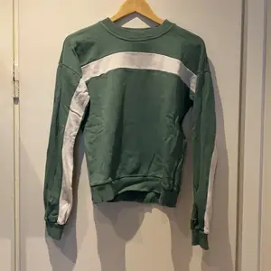 Grön sweatshirt med vitt streck över bröstet som går ut på ärmarna både på fram-och baksidan. Fungerar bra både i sig själv eller med linne under. 