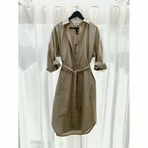 Beige skjort-klänning från House of Dagmar - Helt ny klänning från märket House of Dagmar. 100% organiskt bomull.   Klänning är ljusbeige och har fickor samt skärp. Strl 38.  Passar strl 38 lite oversized och skulle även passa strl 40/42 för en mindre oversized look.   Nypris: ca 3000 kr  Mitt pris: 1800 kr