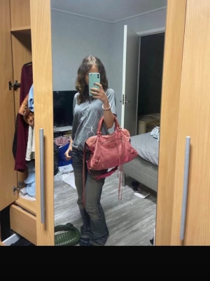 Rosa Balenciaga handväska i skinn - Säljer en snygg rosa handväska från Balenciaga i mjukt skinn. Väskan har coola detaljer med snörning på handtaget, dragkedja framtill och långa remmar. Perfekt storlek för att få plats med allt du behöver och har både handtag och axelrem. Spegel finns och är hel. Lite slitage på handtagen och lite missfärgning men inget som märks av alls. 