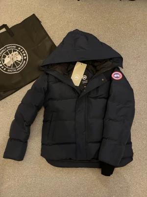 Mörkblå pufferjacka från Canada Goose - Säljer en mörkblå pufferjacka från Canada Goose med huva och klassisk logga på ärmen. Jackan har dragkedja och knappar framtill, samt ribbade muddar vid ärmsluten. Perfekt för kalla vinterdagar och riktigt snygg streetstil.