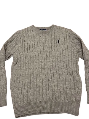 Grå kabelstickad tröja Polo Ralph Lauren - Kabelstickad grå tröja från Polo Ralph Lauren med klassisk rund hals och broderad logga på bröstet. Tröjan har ribbade muddar vid ärmslut och nederkant, och är tillverkad i mjuk bomull. Perfekt för dig som gillar stilrena och tidlösa plagg.