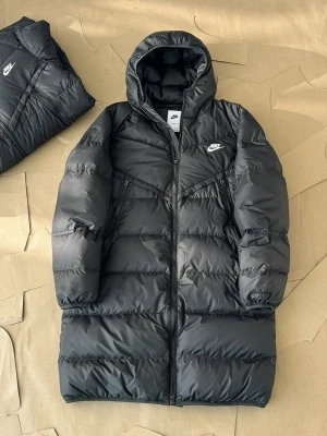  Nike lång dunjacka  - Snygg och varm Nike dunjacka i svart, perfekt för kalla höst- och vinterdagar ❄️ Sportig men stilren design som funkar lika bra till vardags som till träning.  ✨ Detaljer: 	•	Märke: Nike ✔️ 	•	Färg: Svart 	•	Lång modell – extra värme 🔥 	•	Vadderad/pufferjacka 	•	Huva för skydd mot kyla & vind 🧥 	•	Dragkedja framtill 	•	Sidofickor med dragkedja 🔒 	•	Nike-logga på bröstet  📏 Skick: Mycket bra skick 👕 Passform: Normal 