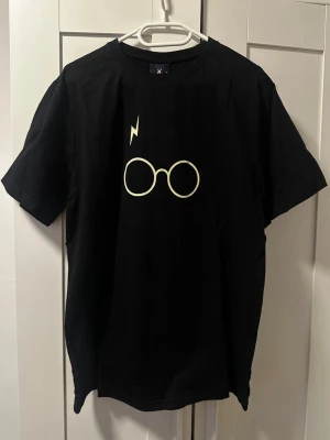 Harry Potter t-shirt från Black Ghost - Svart t-shirt från Black Ghost i bomull med ikoniskt Harry Potter-tryck framtill med glasögon och blixt, samt stor Hogwarts-design och texten 'Harry Potter' på ryggen. Klassisk passform och rund hals. Perfekt för alla Potterheads!