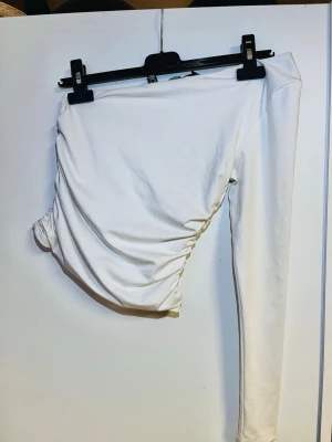 Vit oneshoulder topp från Vero Moda - Säljer en stilren vit oneshoulder topp från Vero Moda i stretchigt material. Toppen har lång ärm på ena sidan och snygg rynkning i sidan för en cool look. Perfekt att matcha med höga jeans eller kjol.