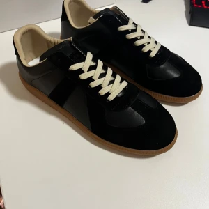 Maison Margiela Gats - 👟 Maison Margiela sneakers i klassisk och tidlös design 🖤 Svart läder med rena linjer och minimalistisk estetik 🤍 Krämfärgad innerfodring och skosnören för elegant kontrast 🟤 Gummisula i naturfärg med bra grepp och hållbar känsla 🔍 Diskret Margiela-detalj baktill som signum för varumärket 📦 Originalförpackning medföljer, inklusive dustbag och box ✨ Mycket fint skick, stilrena och lättmatchade skor