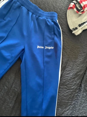 Blåa Palm Angels trackpants M - Säljer ett par blåa trackpants från Palm Angels i storlek M. Byxorna har vita sidoränder, resår i midjan och dragkedjor vid bensluten. Tillverkade i 100% polyester och har Palm Angels-loggan broderad på benet. Perfekta för en sportig och trendig look. Defekter på bilden