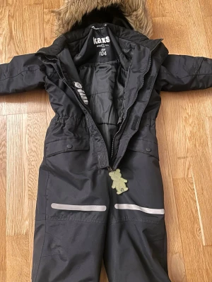 Svarta täckbyxor Kaxs Proxtec strl 104 - Svarta täckbyxor från Kaxs Proxtec med avtagbar huva med fuskpäls, reflexdetaljer på benen och dragkedja framtill. Byxorna har elastisk midja och praktiska fickor med knappar. Perfekta för kalla dagar utomhus.            Ny pris ca 1000kr.                    Pris kan diskuteras