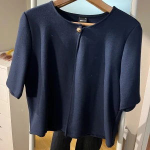 Mörkblå tröja från Gina Tricot - Stilren mörkblå tröja från Gina Tricot med rund halsringning och en dekorativ knapp framtill. Koftan har korta ärmar och ger en modern look. Perfekt att slänga över en topp för extra lager. Knappt använd.