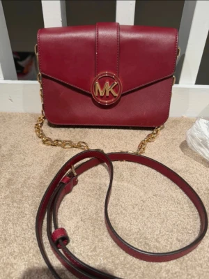 Vinröd axelväska från Michael Kors - Snygg vinröd axelväska från Michael Kors i skinn med guldfärgade detaljer och kedja. Väskan har ett stort MK-emblem framtill, justerbar axelrem och innerfack med dragkedja. Perfekt storlek för det viktigaste och ger en lyxig touch till din outfit.