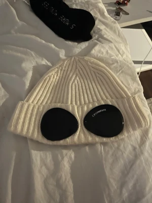 Vit ribbstickad mössa från C.P. Company - Vit mössa från C.P. Company. Mössan är i grovstickad ullblandning. Perfekt accessoar för att sticka ut i vinterkylan. (OBS finns pytte lite sminkfläckar i mössan efter att min syster använt den 1 gång men går och få bort) annars utmärkt skick. Pris går o diskutera vid snabb affär 