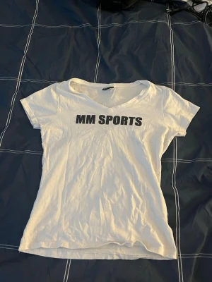 Vit t-shirt från MM Sports - Säljer en enkel och clean vit t-shirt från MM Sports med svart tryck på bröstet. T-shirten har v-ringad hals och korta ärmar, perfekt för dig som gillar sportig och avslappnad stil. Vet ej om det är dam eller herr modell.