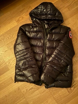 Canada goose crofton  - Säljer en svart, glansig dunjacka från Canada Goose med huva och klassisk patch på ärmen. Jackan har dragkedja framtill och är quiltad i breda paneler. Perfekt för kalla dagar och riktigt snygg till streetwear-stilen.