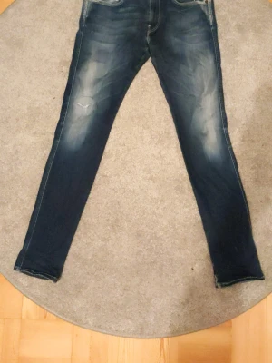 Replay Anbass jeans blå, storlek 32 - Säljer ett par Replay Anbass jeans i mörkblå tvätt med slitningar och detaljer på benen. Klassisk femficksmodell med raka ben och kontrastsömmar. Jeansen är tillverkade i mjukt denimtyg och har en modern passform. Det fanns ett hål vid ett av knäna men det är igensytt nu