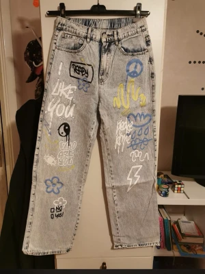 Grå jeans med graffiti-tryck - Unika grå jeans med streetinspirerade graffiti-tryck i vitt, blått och gult. Rak passform och klassisk femficksmodell. Trycken inkluderar blommor, peace-märke, blixt och olika texter. Perfekta för dig som vill sticka ut med en kreativ och personlig stil. Använde fåtal gånger. Välj helst insta box då både du och jag slipper fraktkostnaden!💛