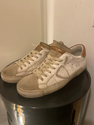 Philippe Model vita sneakers - Säljer ett par vita Philippe Model sneakers med beige mockadetaljer och bruna skinnpartier vid hälen. Skorna har snörning, rund tå och platt sula. Klassisk och stilren design som passar till det mesta. Perfekta för dig som gillar en avslappnad men ändå snygg look.