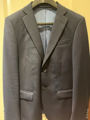 Mörkblå kavaj Dressmann slim fit - Snygg mörkblå kavaj från Dressmann i slim fit-modell. Klassisk design med två knappar, smala slag och innerfoder i matchande blått. Perfekt för dig som vill ha en stilren och modern look. Storlek 48 Short.