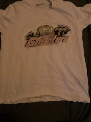 Vit Trapstar London Shooters t-shirt - Säljer en vit t-shirt från Trapstar med trycket 'London Shooters' och coolt motiv på bröstet. Klassisk passform och rund hals. Perfekt för dig som gillar streetwear och vill sticka ut med en unik design.