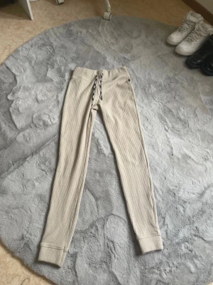 Beige ribbade byxor från Stronger - Säljer ett par beige ribbade byxor från Stronger i storlek small. Byxorna har hög midja, svart snörning med logga och är tillverkade i ett mjukt, stretchigt material. Perfekta för chill eller träning och riktigt sköna att ha på sig.