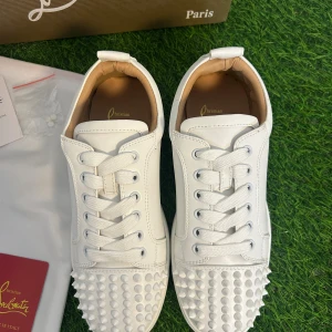 Christian Louboutin vita sneakers med nitar - Säljer ett par sjukt snygga vita sneakers från Christian Louboutin med ikonisk röd sula och coola nitar på tån. Skorna har snörning, rund tå och är tillverkade i lyxigt skinn. Perfekt för dig som vill sticka ut med exklusiv stil och detaljer. Använd ca 3-4 gånger