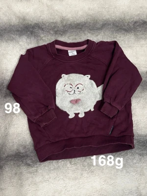 Vinröd sweatshirt med kattmotiv - Mysig vinröd sweatshirt med ett fluffigt kattmotiv framtill. Tröjan har långa ärmar, ribbade muddar och rund hals. Perfekt för dig som gillar gulliga detaljer och vill ha något bekvämt till vardags.🌸 Fint använt skick. Storlek 98