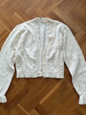 Vit stickad kofta med knappar - Mysig vit stickad med dekorativt hålmönster och ribbade muddar. Koftan har v-ringning, långa ärmar och stängs med fem knappar framtill. 