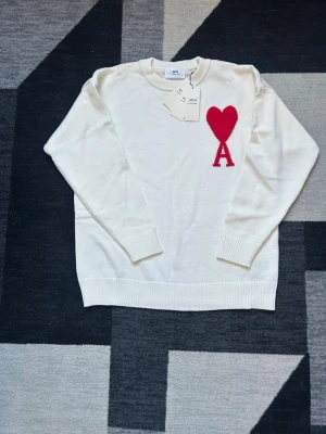 Vit stickad tröja från Ami Paris - Säljer en vit stickad tröja från Ami Paris med röd hjärtdetalj och bokstaven A på bröstet. Tröjan har rund halsringning, ribbade muddar och är långärmad. Perfekt för dig som vill ha en clean look med en twist. Kan diskutera pris 