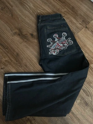 JNCO jeans - Heans från JNCO med broderad krona och graffiti på bakfickan. Snygga kontrastsömmar och vita stripes längs benen ger en retro vibe. 35 cm i platt midjemått.
