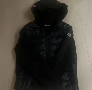 Svart Moncler dunjacka med huva - Svart dunjacka från Moncler med glansig yta och huva. Jackan har Moncler-logga på ärmen, dragkedja framtill och är quiltad på kroppen. Fylld med vitt gåsdun enligt DIST-standard. Perfekt för dig som vill ha en snygg och varm jacka med street-vibe.