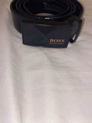 Äkta Hugo Boss bälte i skinn - Snyggt svart bälte från Hugo Boss med stilren metallspänne där loggan syns i guld. Bältet är gjort i slätt skinn och har en modern, minimalistisk look som passar till många outfits. Perfekt accessoar för att lyfta din stil.