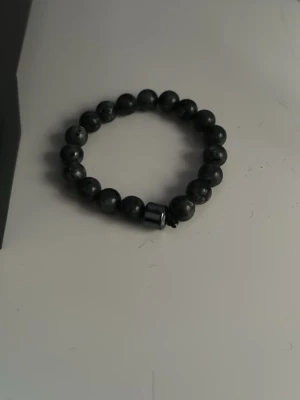 Svart pärlarmband med cylinderpärla - Säljer ett stilrent svart armband med runda pärlor och en större cylinderformad pärla i mitten. Armbandet har en elastisk tråd som gör det enkelt att ta på och av. Perfekt accessoar för dig som gillar minimalistisk och clean stil.