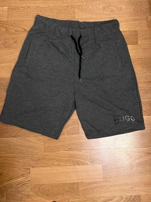 Mörkgrå shorts  - Säljer ett par mörkgrå shorts med svart snörning i midjan och diskret logga på benet. De har en avslappnad passform och är tillverkade i mjuk bomull, perfekta för chill dagar eller träning.