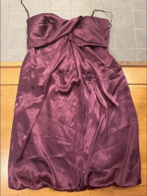 Vinröd axelbandslös kortklänning - Säljer en vinröd axelbandslös kortklänning i glansigt material med draperad överdel och lätt omlottkänsla. Perfekt för dig som vill ha en elegant look med en twist. Klänningen har tunna axelband och en mjuk, följsam passform.