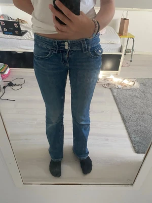 Blå bootcut jeans med låg midja - Säljer ett par blå jeans med bootcut passform och låg midja. Jeansen har dubbla knappar i linningen, klassiska fickor fram och bak samt snygga sömdetaljer. Materialet är jeans i bomull och de har en lätt utsvängd siluett nedtill. Storlek 152 men stora i storleken.