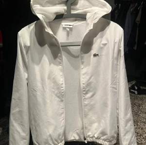 Vit vindjacka från Lacoste med luva - Pris kan diskuteras ! Snygg vit vindjacka från Lacoste med klassisk krokodillogga på bröstet och tryckt Lacoste-text på ärmen. Jackan har huva, dragkedja framtill och elastisk nederkant. Materialet är lätt och syntetiskt, perfekt för blåsiga dagar.
