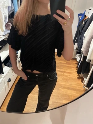 Svart topp - Säljer en svart croppad topp med diagonala ränder med fransar. Toppen har korta ärmar och en lös passform som ger en avslappnad vibe. Materialet känns mjukt och strukturerat, perfekt till jeans eller kjol. Toppen går att ha off shoulder