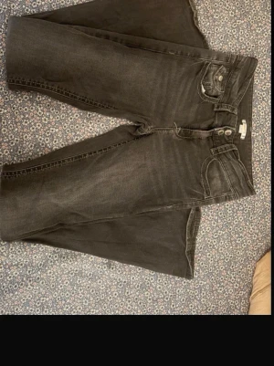 Svarta bootcut jeans med dubbla knappar - Säljer ett par svarta bootcut jeans med dubbla knappar i midjan och klassiska fem fickor. Lågmidjade jeans i gråsvart färg. Perfekta för dig som gillar en avslappnad men ändå snygg stil.
