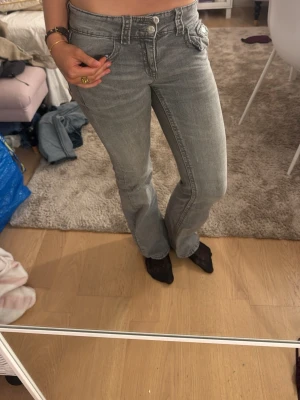 Grå bootcut jeans med dubbla knappar - Säljer ett par grå jeans med bootcut passform och dubbla knappar i midjan. Jeansen har klassiska fickor fram och bak, och är tillverkade i ett mjukt jeansmaterial.