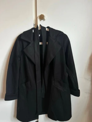 Svart kappa med bälte - Stilren svart kappa med knytbälte i midjan och klassisk krage. Jackan har långa ärmar och en rak passform som ger en snygg siluett. Perfekt för kyligare dagar och enkel att matcha med olika outfits.  Den är helt ny och oanvänd därav priset. Nypris:600kr!