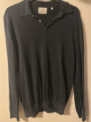 Svart pikétröja från Bläck - Klassisk mörkblå pikétröja från Bläck med lång ärm och mjuk krage. Tröjan har två knappar vid halsen och ribbade muddar vid ärmslut och nederkant. Perfekt till en clean och enkel stil.