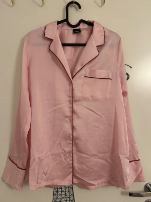 Ljusrosa pyjamasskjorta i satin från Gina Tricot - Ljusrosa pyjamasskjorta i satin från Gina Tricot i storlek S med röda kontrastkanter, klassisk krage och bröstficka. Skjortan har långa ärmar och har knäppning framtill. Skjortan är i nytt skick då den aldrig är använd. 