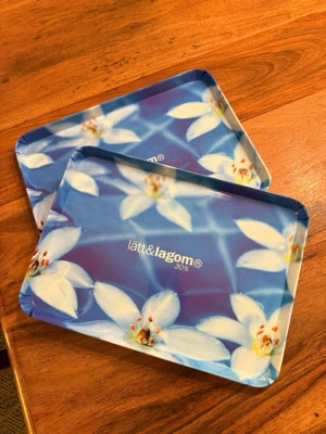 Blå brickor med blommigt mönster – Lätt & Lagom - Säljer två snygga brickor i blått med blommigt mönster från Lätt & Lagom. Perfekta för servering eller som dekor i köket. I fint skick.  Mått: 30 × 22 cm