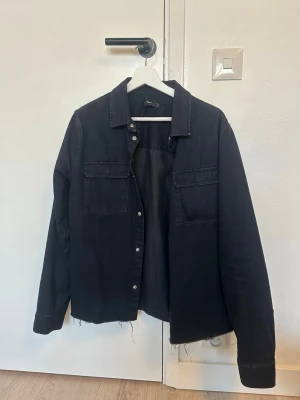 Svart jeansjacka med rå kant - Säljer en svart jeansjacka med rå, fransig nederkant och klassisk krage. Jackan har två bröstfickor med lock och knäppning framtill. Perfekt för dig som gillar en avslappnad och trendig stil.