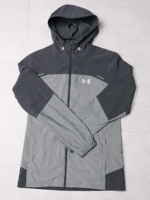 Under armour jacka  - Snygg tvåfärgad vindjacka från Under Armour i mörkgrått och ljusgrått. Jackan har huva, dragkedja framtill och logga på bröstet. Perfekt för blåsiga dagar och har en sportig look med långa ärmar och praktiska detaljer.