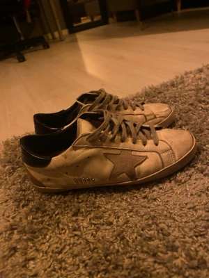 Vita sneakers med stjärndetalj - Säljer ett par vita sneakers från Golden Goose med ikonisk stjärna på sidan, svarta detaljer vid hälen och grå snörning. Skorna har en platt sula och är tillverkade i skinn och mocka för en cool, avslappnad look.