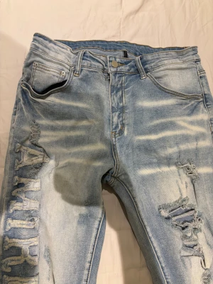 Ljusblå ripped jeans från Amiri - Säljer ett par ljusblå skinny jeans från Amiri med slitna detaljer och stora ripped hål på benen. Snyggt broderat AMIRI-tryck längs vänster ben och klassisk femficksmodell. Jeansen har en tvättad look och är tillverkade i bomull med stretch. Storlek S